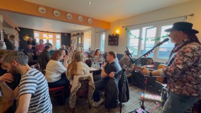 Wiggy singing and playing acoustic guitar to a seated pub audience